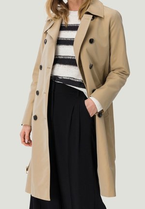 Kobieta w beżowym, dwurzędowym płaszczu trencz na czarnych spodniach i czarno-białym pasiastym swetrze, jedna ręka w kieszeni.