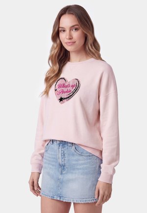 Donna con capelli lunghi e mossi indossa una felpa rosa chiaro con un cuore e testo "What's up Pookie" e una minigonna di jeans azzurro chiaro.
