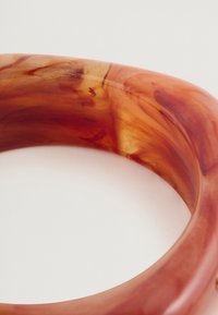 Διαυγές πορτοκαλί-κόκκινο ακρυλικό βραχιόλι με στριφτούς σχεδιασμούς, λεία υφή και στρογγυλό σχήμα, διαθέτοντας γυαλιστερή επιφάνεια.