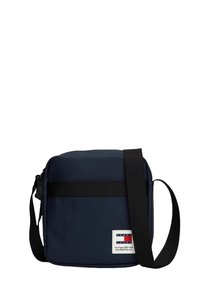 Granatowa torebka na ramię z gładką teksturą, wyposażona w czarny pasek, białą metkę marki oraz prostokątny kształt z zaokrąglonymi krawędziami.