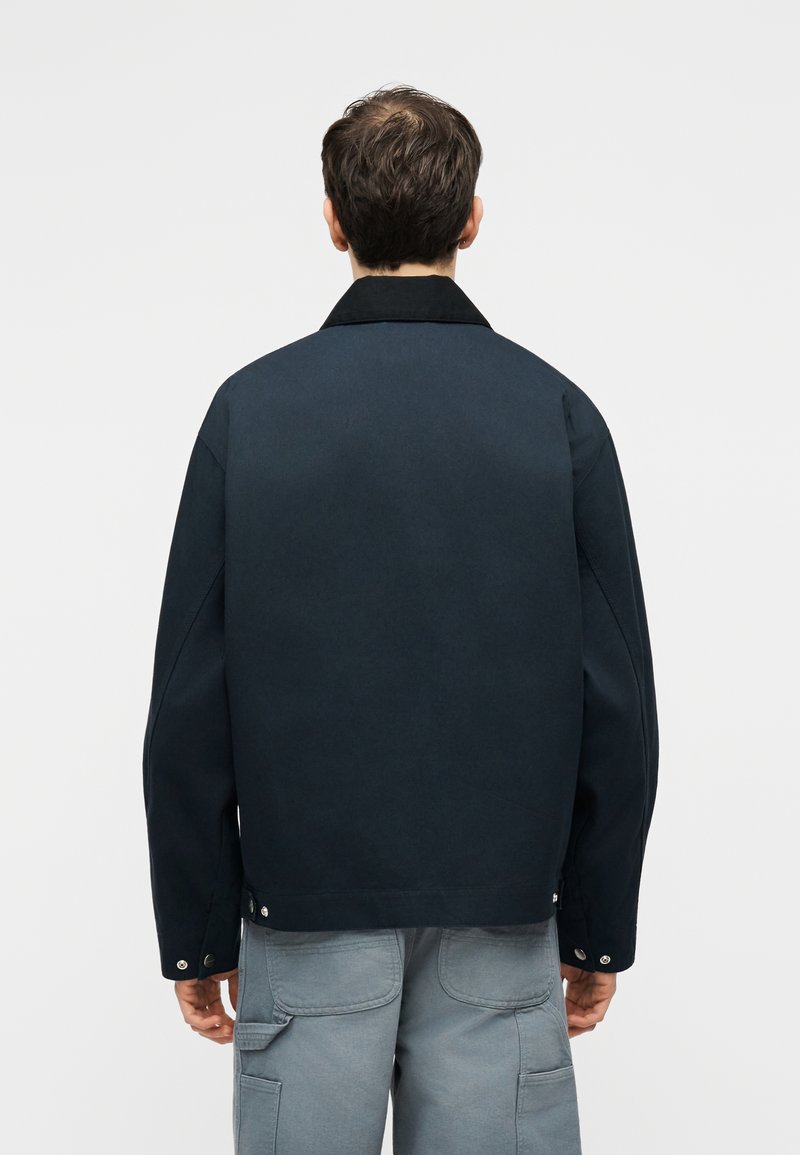 Man met een donkerblauw jack en lichtgrijze cargobroek, staand met zijn rug naar de kijker tegen een effen lichte achtergrond.