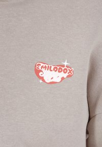Tissu de sweat-shirt beige clair présentant un logo rouge et blanc proéminent avec le texte « EMILODOX » et des accents d'étoiles décoratifs.