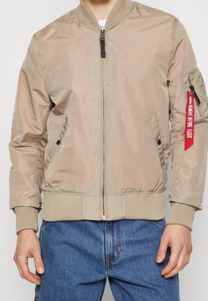 Beige bomberjakke med ribstrikkede manchetter og kant, lynlås foran, sidelommer og en rød "Remove Before Flight"-mærkat på venstre ærmelomme.