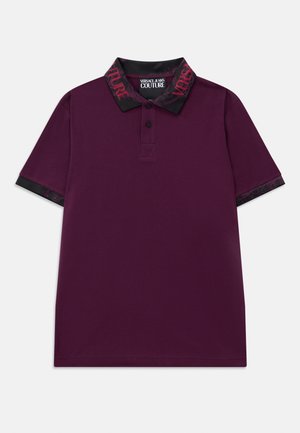 Violets īsām piedurknēm polo krekls ar melnu apkakli un piedurknēm, uz kura ir sarkans teksts "Versace Jeans Couture" un divas priekšējās pogas.