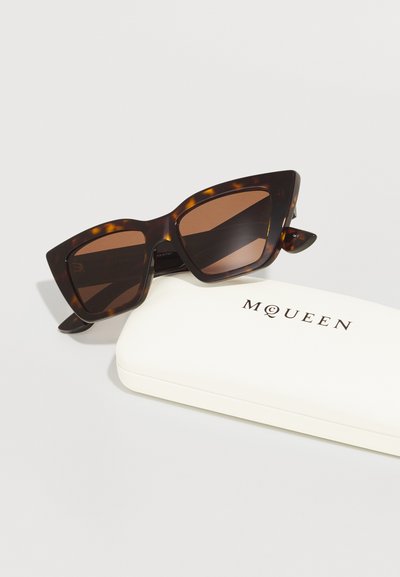 Cat-eye sunglasses in tortoiseshell acetate with brown lenses, paired with a cream-colored hard case featuring the brand name "MQUEEN."