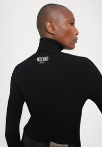Svart ribbestrikket turtleneck-genser med en tettsittende design, høy hals og et Moschino Jeans logo-patch på ryggen.