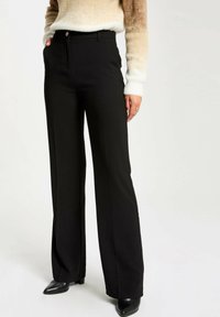 Pantalones negros ajustados con una cintura entallada, piernas acampanadas y una textura suave. Presentan un cierre delantero de cremallera y botón, combinados con tacones negros.