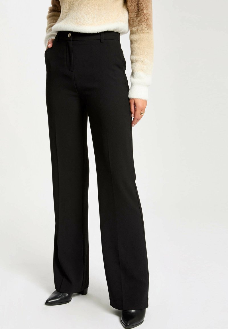 Pantalones negros ajustados con una cintura entallada, piernas acampanadas y una textura suave. Presentan un cierre delantero de cremallera y botón, combinados con tacones negros.