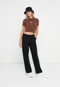 Brun cropped t-skjorte med logo, svarte løse bukser og hvite sneakers. Modellen har på seg en svart bøttehatt. Glatte teksturer og avslappet stil.