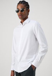 White long-sleeve shirt made of smooth fabric, featuring a half-open collar and button closure, worn with round sunglasses against a neutral background.