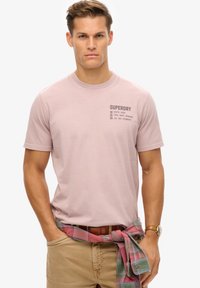 Lyserød kortærmet t-shirt med "Superdry" logo i grå på fronten. 100% bomuldsmateriale, standard pasform, parret med khaki bukser og en ternet skjorte bundet om taljen.
