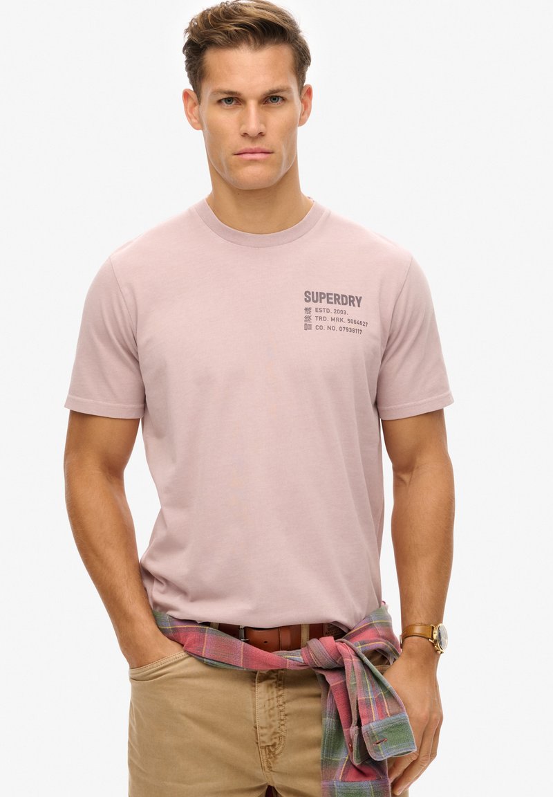 Lyserød kortærmet t-shirt med "Superdry" logo i grå på fronten. 100% bomuldsmateriale, standard pasform, parret med khaki bukser og en ternet skjorte bundet om taljen.