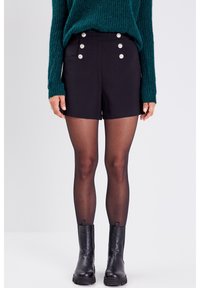 Shorts negros a medida con bolsillos laterales y botones plateados decorativos. Llevados con botas negras hasta el tobillo y medias oscuras. Suéter de punto verde por encima.