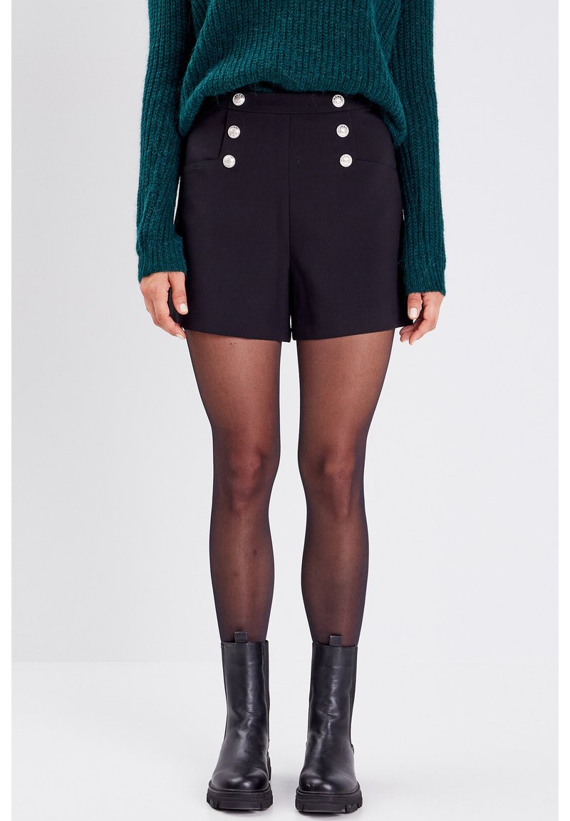 Shorts negros a medida con bolsillos laterales y botones plateados decorativos. Llevados con botas negras hasta el tobillo y medias oscuras. Suéter de punto verde por encima.