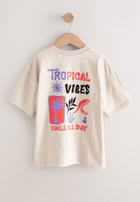 Beige short-sleeve t-shirt hanging on wooden hanger with colorful text "Tropical Vibes Chill All Day" and tropical-themed illustrations.