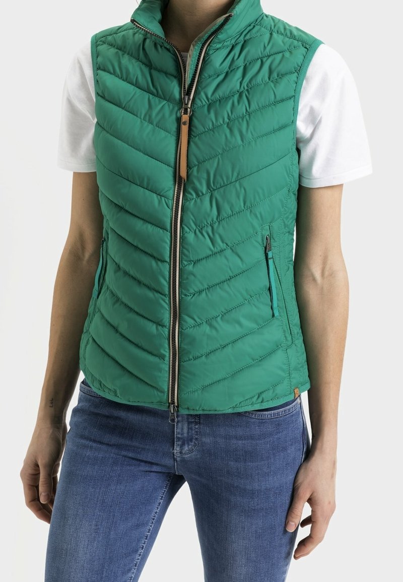 Veste matelassée verte avec col montant, fermeture éclair avant et languette en cuir. Dotée de poches latérales et d'une coupe ajustée, portée avec une chemise blanche.