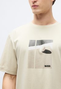 Beżowy bawełniany t-shirt z nadrukiem przedstawiającym piłkę na piaszczystej powierzchni, z czarnym logo "BOSS" w prawym dolnym rogu.