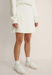 Valkoinen plisattu minihame, yhdistetty valkoiseen ylisuureen huppariin ja valkoisiin lenkkareihin. Hameessa on sileä rakenne ja hyvin istuva vyötärö.