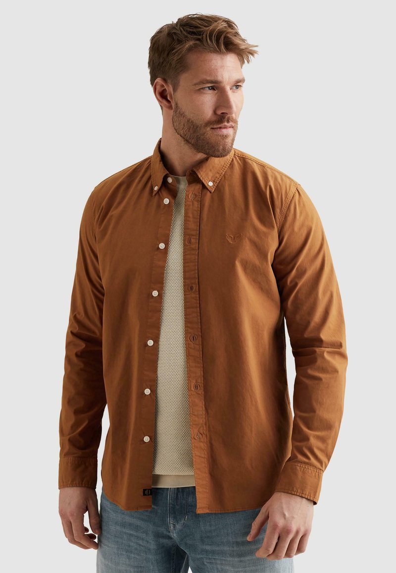 Homme avec une barbe châtain clair portant une chemise couleur rouille déboutonnée par-dessus un pull beige et un jean bleu, regardant vers la droite.