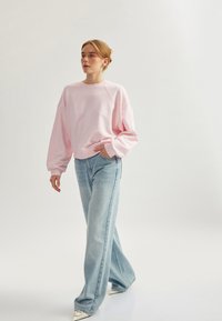 Felpa oversized rosa chiaro con maniche a raglan, abbinata a jeans larghi blu sbiaditi e scarpe argento a punta, su uno sfondo neutro.