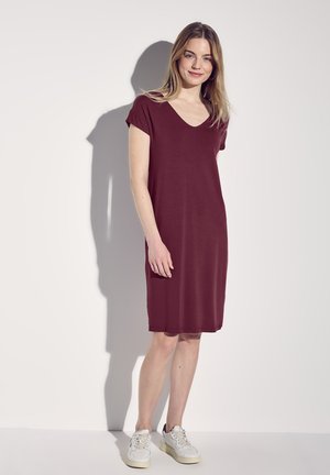 Robe bordeaux à manches courtes avec un col en V, d'une longueur au genou, fabriquée en tissu lisse. Associée à des baskets blanches.