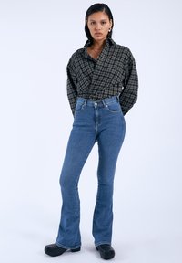 Camisa envolvente de cuadros en blanco y negro con un alto cuello, combinada con jeans acampanados de tiro alto en azul y zuecos negros. Textura de tela suave.