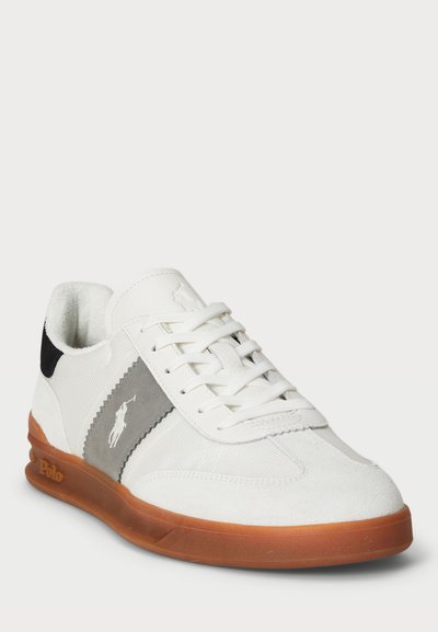 Sapatilha desportiva branca com detalhes cinzentos, tecido texturizado e sola de borracha, apresentando um logótipo e um design com atacadores. A sola castanha melhora a aderência.