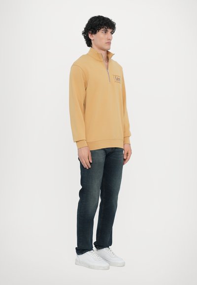 Sweat à zip couleur moutarde avec des manches longues, arborant un logo sur la poitrine. Associé à un jean sombre et des baskets blanches.