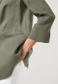 Olivgrüner Leinenblazer mit einer einzelnen Tasche und umgeschlagenen Ärmelbündchen, der eine lockere Passform und seitliche Schlitze für mehr Bewegungsfreiheit bietet.