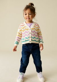 Gebreid vest met een crèmewitte basis, voorzien van golvende, meerkleurige strepen in geel, roze en groen. Gecombineerd met donkere spijkerbroek en patroon schoenen.