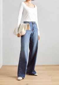 Kvinna som bär en vit långärmad topp, högmidjade vida blå jeans, vita skor och håller en beige hopvikt jacka mot en enfärgad vägg. 