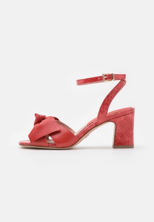 Sandale rouge à bout ouvert avec détail nœud, bride de cheville avec boucle dorée, et talon bloc avec motif texturé subtil.