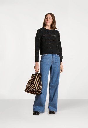 Femme portant un pull en maille noir et un jean large bleu, tenant un grand sac fourre-tout rayé noir et marron, debout devant un fond blanc.
