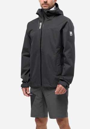 Mann trägt schwarze wasserdichte Jacke mit Kapuze und graue Outdoor-Shorts vor einem einfarbigen hellen Hintergrund.