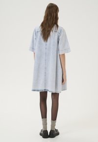 Robe en denim bleu clair à manches courtes, coupe décontractée, ourlet effiloché et texture douce. Portée sur des collants noirs foncés et des chaussures noires imposantes.