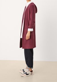 Lunga cardigan lavorata a maglia fucsia con cappuccio, texture a coste, tasche laterali, indossata sopra una camicia bianca, abbinata a jeans scuri e sneakers.