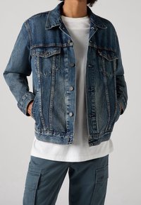 Blå jeansjacka med knäppning, två bröstfickor och en lätt sliten textur, bärs över en vit skjorta och olivgröna byxor.