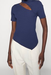 Marinblå ribbad topp med korta ärmar, hög krage och asymmetrisk nederkant. Noterbart utskärningsdetalj på bröstområdet.