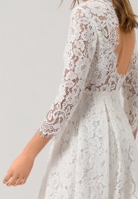 Vit spetsklänning med trekvartsärmar och öppen rygg, bärs av en person som visar detaljerat blommönstrad spets och plisserad kjol.