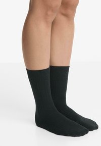 Gambe che indossano calzini neri a costine semplici, in piedi su una superficie bianca con uno sfondo chiaro.