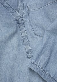 Ljusblå denimskjorta med V-hals, en bröstficka och samlade, elastiska ärmar med synliga sömdetaljer.