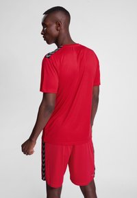Conjunto de camiseta atlética roja y pantalones cortos; hecho de material ligero; presenta acentos en negro en forma de chevrón en los hombros y los lados.