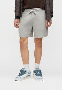 Άνδρας που φοράει ανοιχτό γκρι σορτς με κορδόνι, λευκές κάλτσες μέχρι τον αστράγαλο και μπλε-λευκά αθλητικά παπούτσια, στέκεται μπροστά σε μονόχρωμο φόντο.