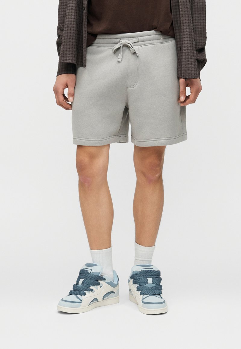 Άνδρας που φοράει ανοιχτό γκρι σορτς με κορδόνι, λευκές κάλτσες μέχρι τον αστράγαλο και μπλε-λευκά αθλητικά παπούτσια, στέκεται μπροστά σε μονόχρωμο φόντο.