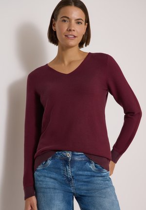 Femme portant un pull bordeaux à manches longues et col en V ainsi qu'un jean bleu, debout devant un fond clair uni.