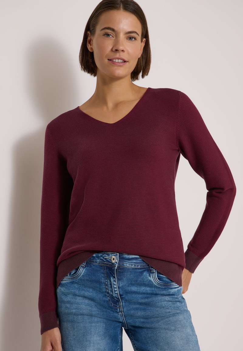 Femme portant un pull bordeaux à manches longues et col en V ainsi qu'un jean bleu, debout devant un fond clair uni.