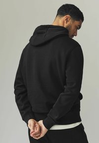 Sudadera negra con capucha, material de mezcla de algodón, puños y dobladillo acanalados, y un ajuste relajado. Bolsillo visible en la cintura.