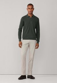Suéter de color verde oscuro con cuello de solapa y tres botones, combinado con pantalones beige claro y zapatos de cuero oscuro, con una textura suave.