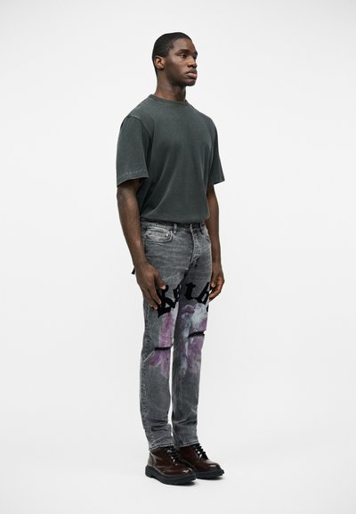 Άνδρας που στέκεται πλαγίως φορώντας σκουρό πράσινο φαρδύ μπλουζάκι, γκρι τζιν με μοτίβα σε μωβ και μαύρο, και καστανά παπούτσια, με φόντο λευκό.