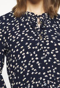Granatowa bluzka z drobnym kwiatowym wzorem w białym i żółtym kolorze. Posiada dekolt ze sznurkiem, zapięcie na guziki oraz teksturowaną tkaninę z lekkim połyskiem.
